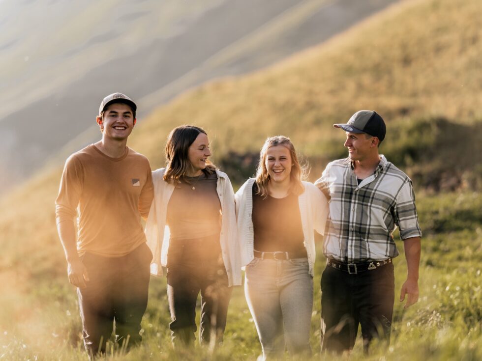 Vier Freunde spazieren lachend durch eine Bergwiese im goldenen Abendlicht